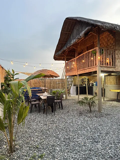 Raised bamboo kubo with a thatched roof above courtyard tables and string lights.